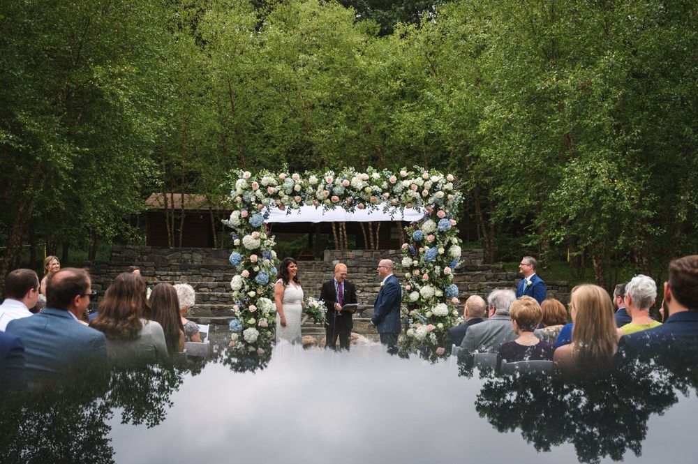 Outdoor wedding ceremony under a floral arch with guests seated on white chairs in a wooded setting.