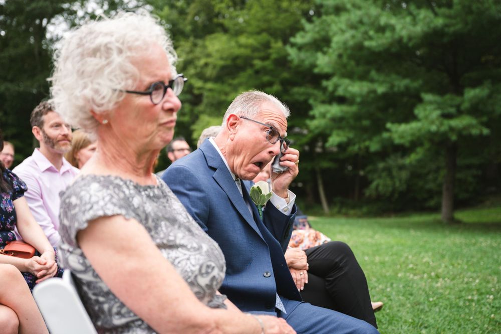 Wedding guests react emotionally during an outdoor ceremony with lush green trees in the background.