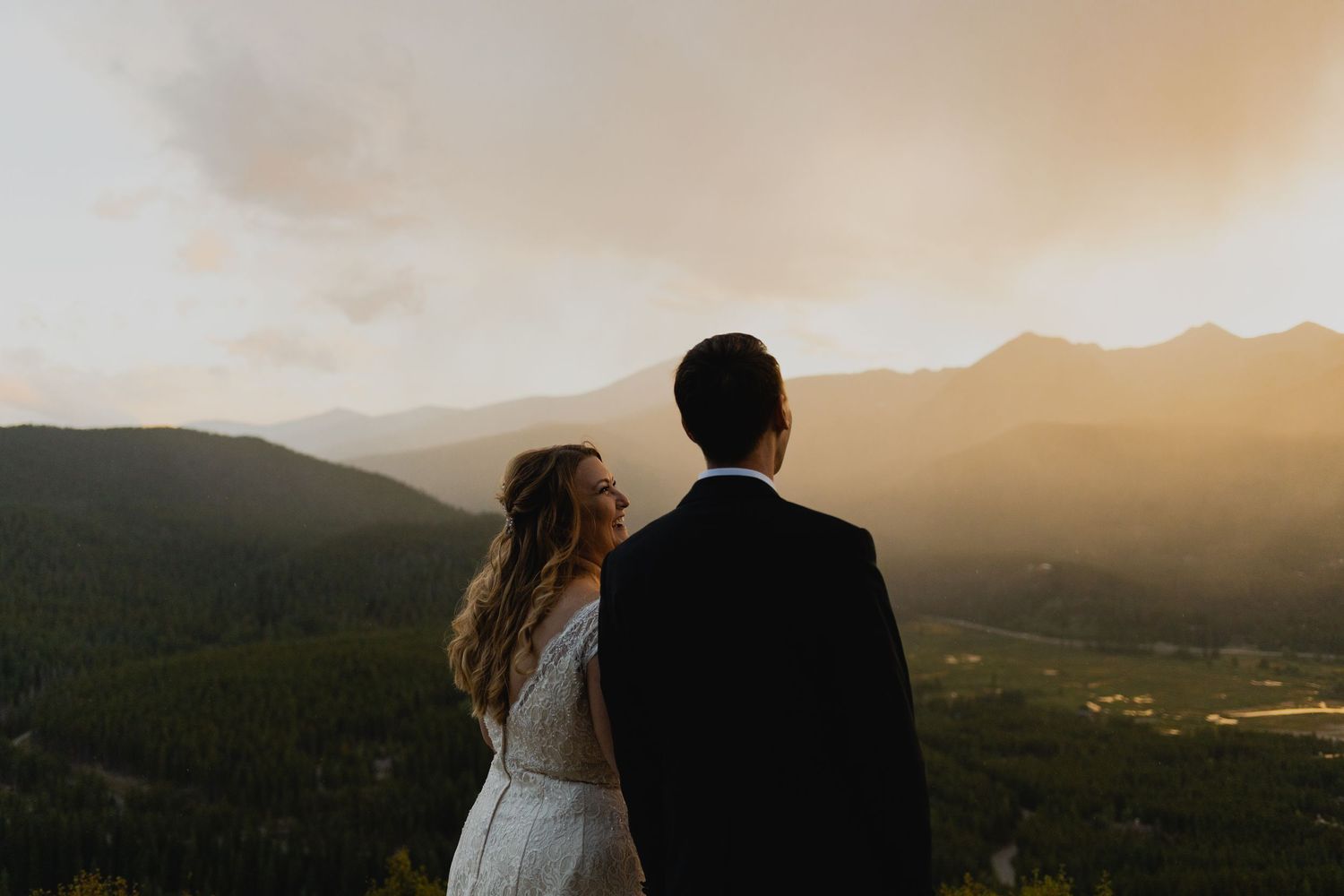Couple in wedding attire stands silhouetted against a dramatic mountain landscape at sunset, overlooking a scenic valley.