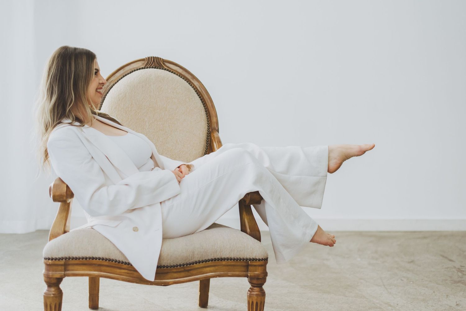 A woman in a white linen suit relaxes sideways on an elegant vintage chair against a light background.