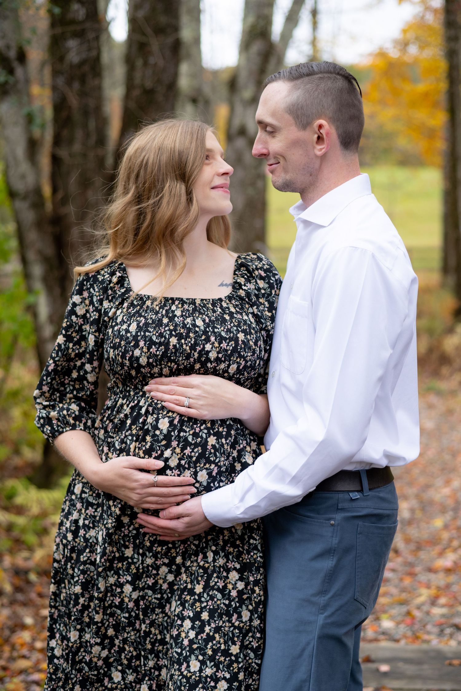 A couple stands lovingly together in an autumn forest setting, with fallen leaves and golden foliage in the background.