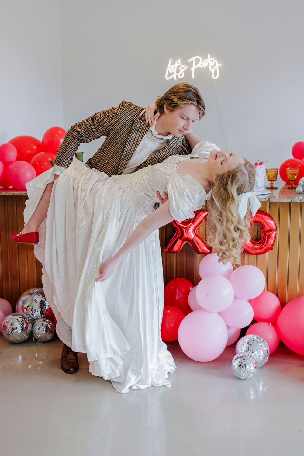 Romantic Valentine's Day photoshoot with couple dancing surrounded by red and pink balloons in decorated room.