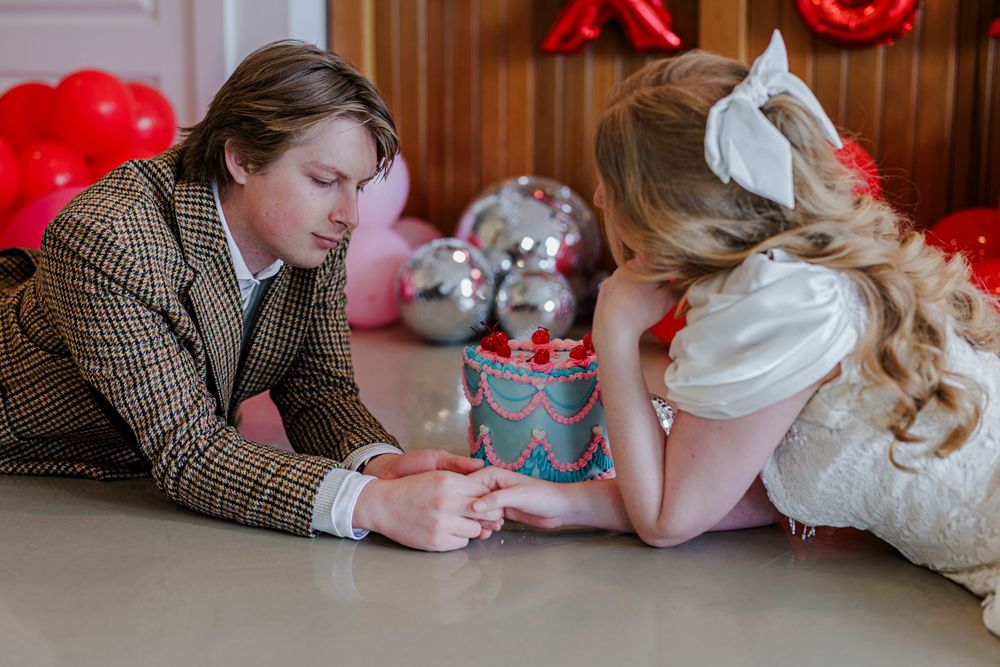 Two people lying on the floor and sharing a Valentine's Day moment with red balloons and heart decorations in the background.