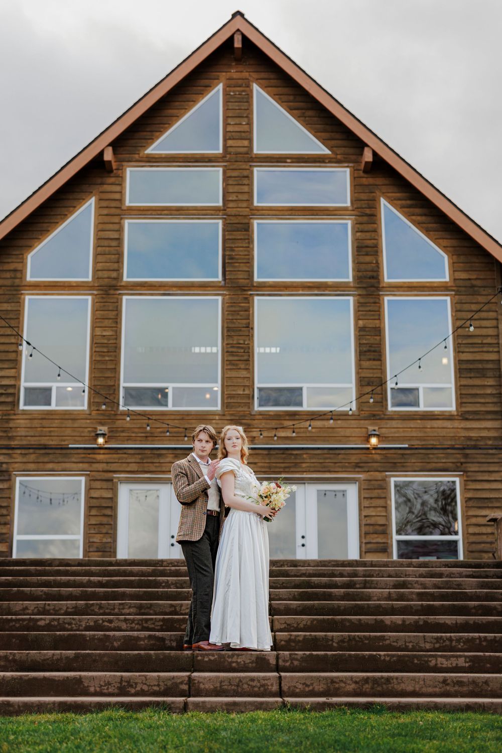 A rustic wooden lodge with large triangular windows serves as backdrop for a romantic moment on stone steps.