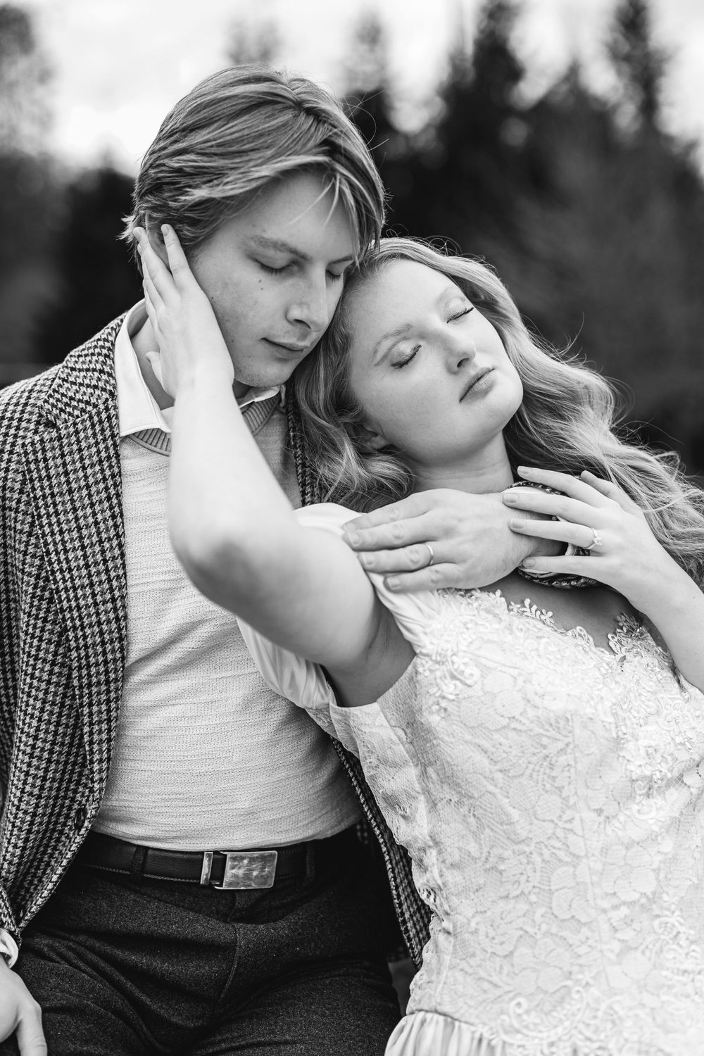 A black and white romantic portrait in front of evergreen trees shows intimate embrace in wedding attire.