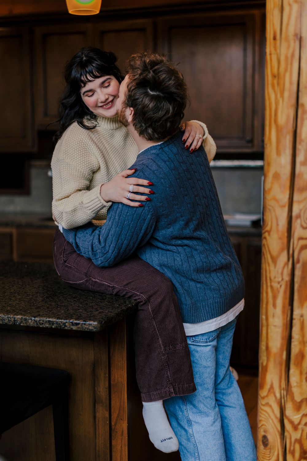 Two people share an intimate moment in a kitchen, wearing cozy sweaters while seated on wooden furniture.