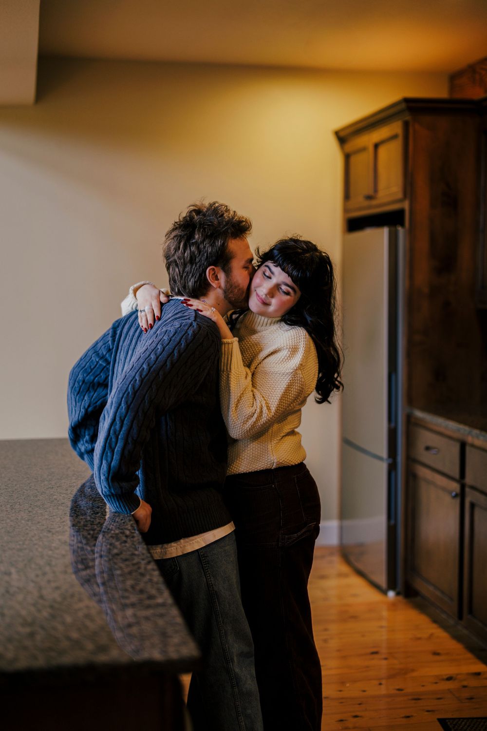 A romantic couple embraces in a cozy kitchen with warm lighting and wooden floors.