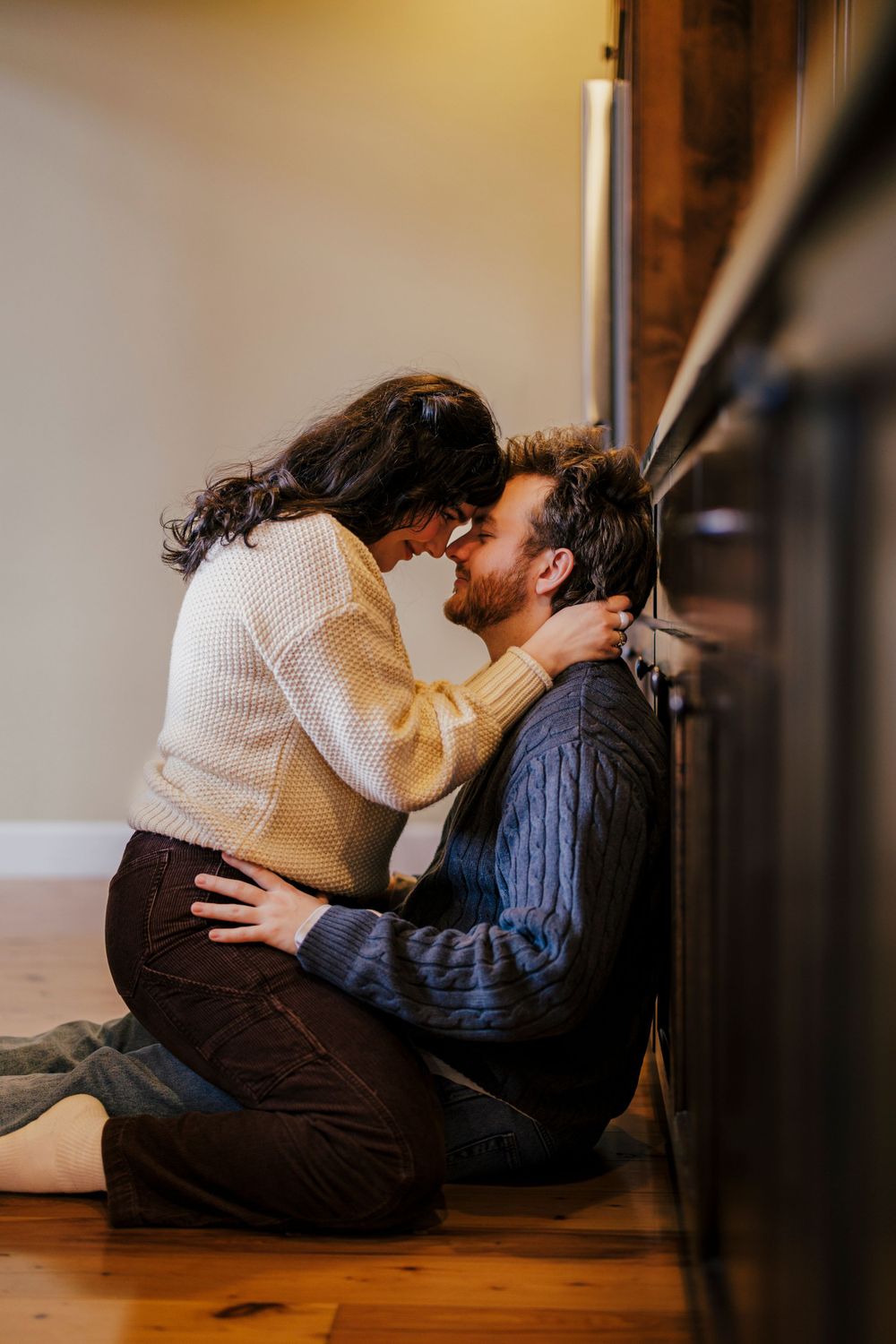 A romantic couple embraces tenderly while sitting on a wooden floor in warm indoor lighting.