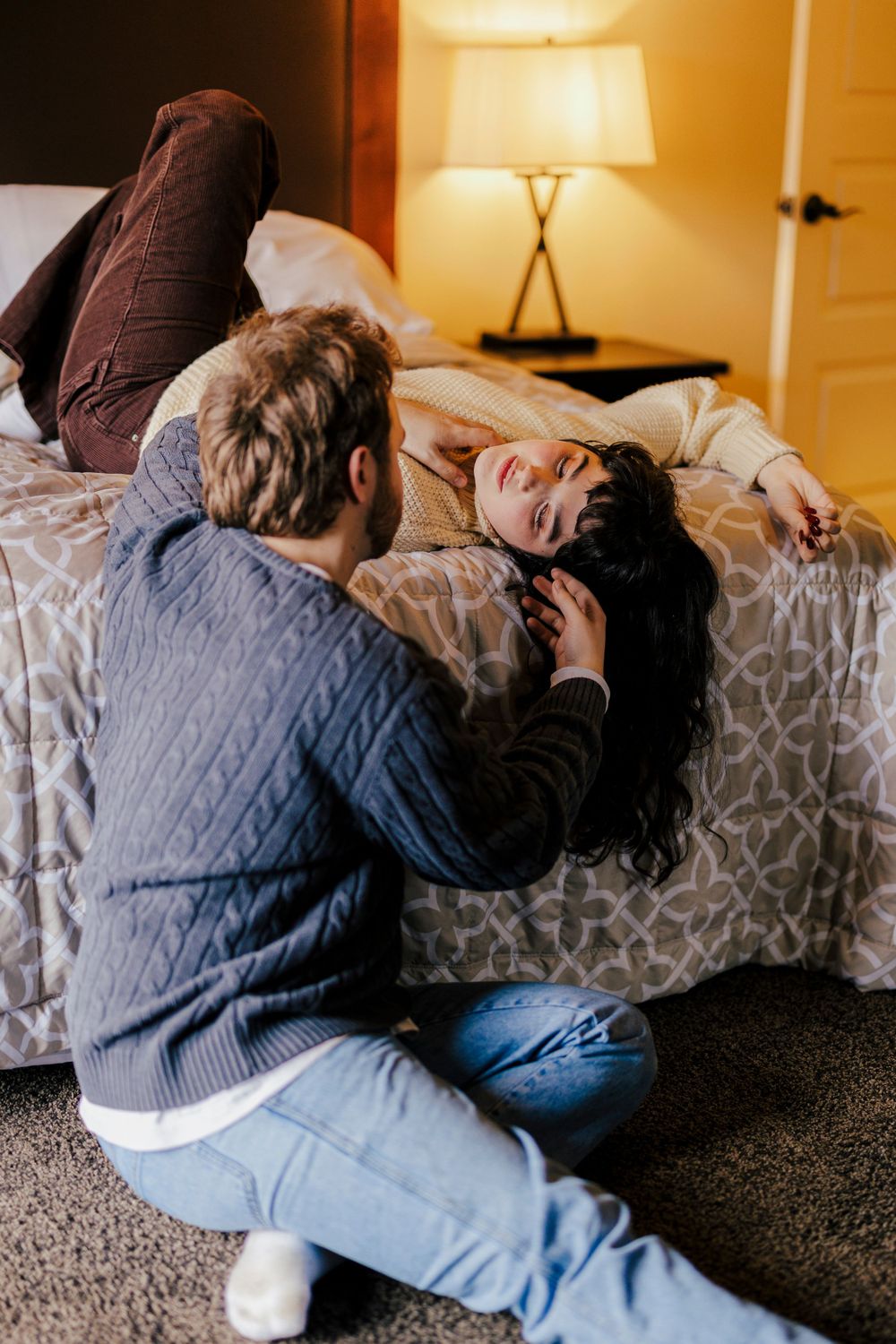 A couple relaxes intimately on a bed in a warmly lit room with a table lamp creating cozy ambiance.