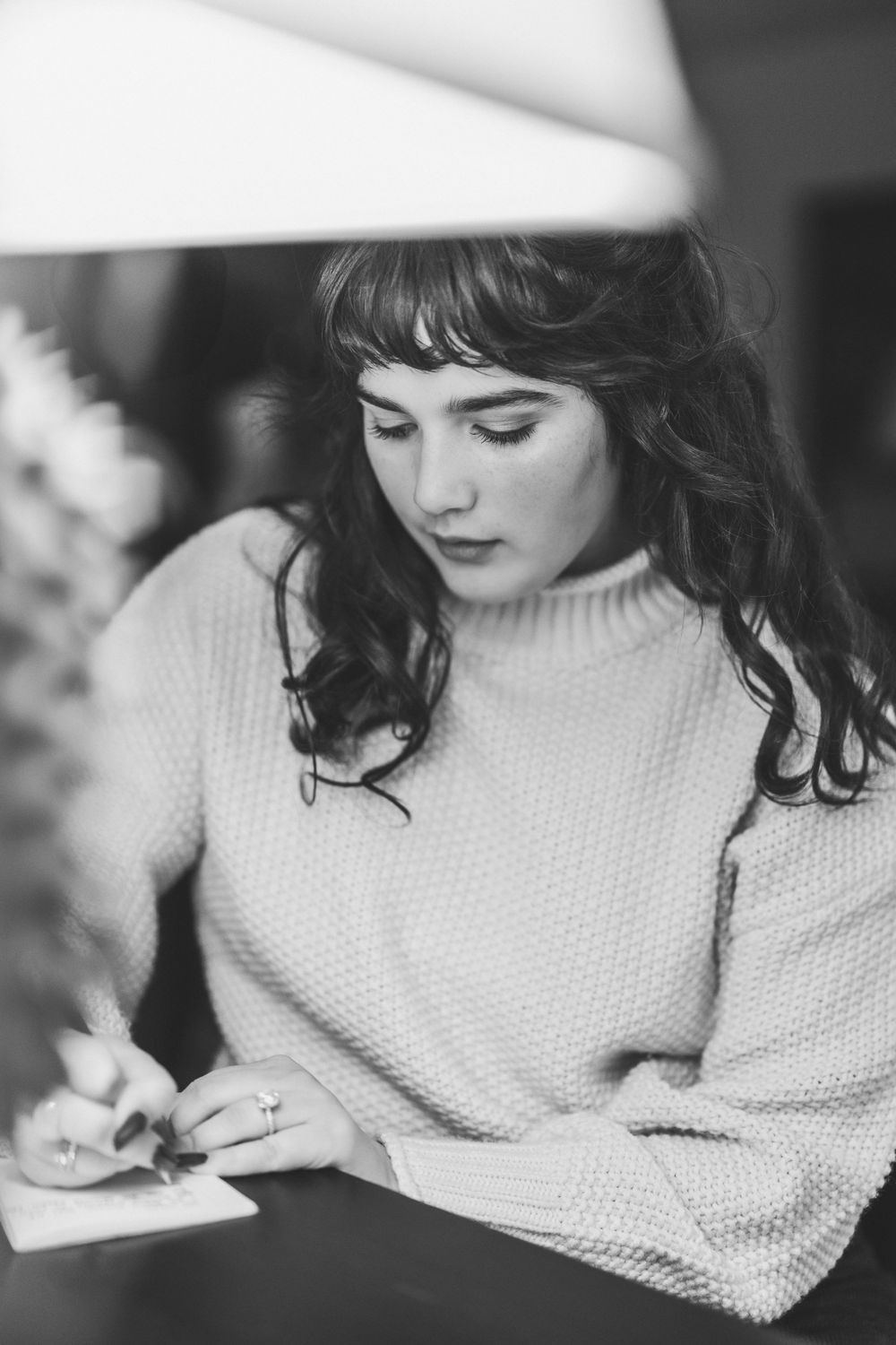 Black and white portrait of a person with dark hair wearing a cozy sweater while writing or drawing at a table.