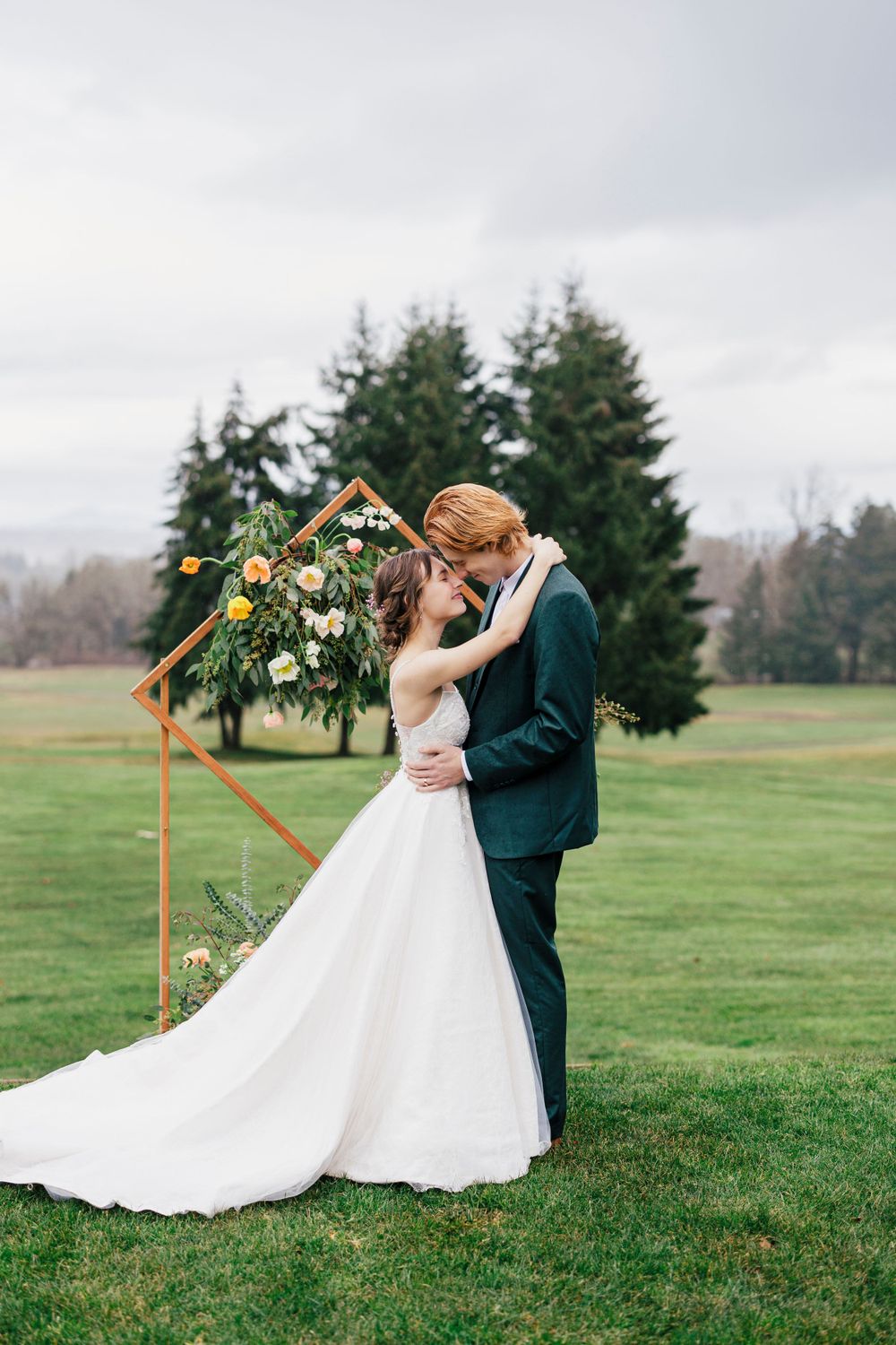 A couple shares a romantic moment under a floral arch on a misty green lawn with evergreen trees in the background.
