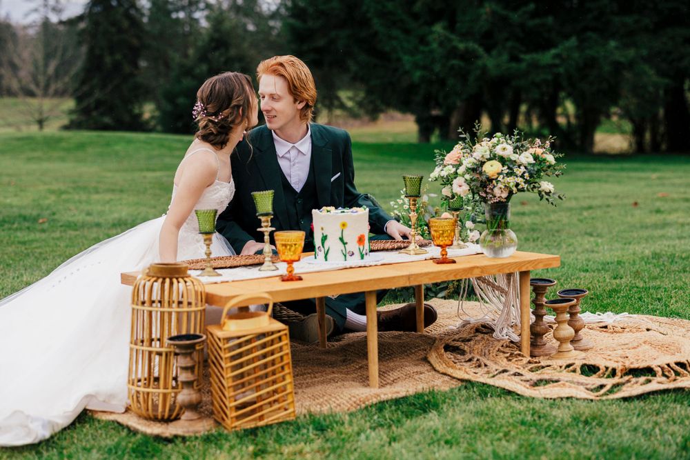 A couple enjoys an intimate wedding picnic at a low wooden table on grass with wine and floral centerpiece.