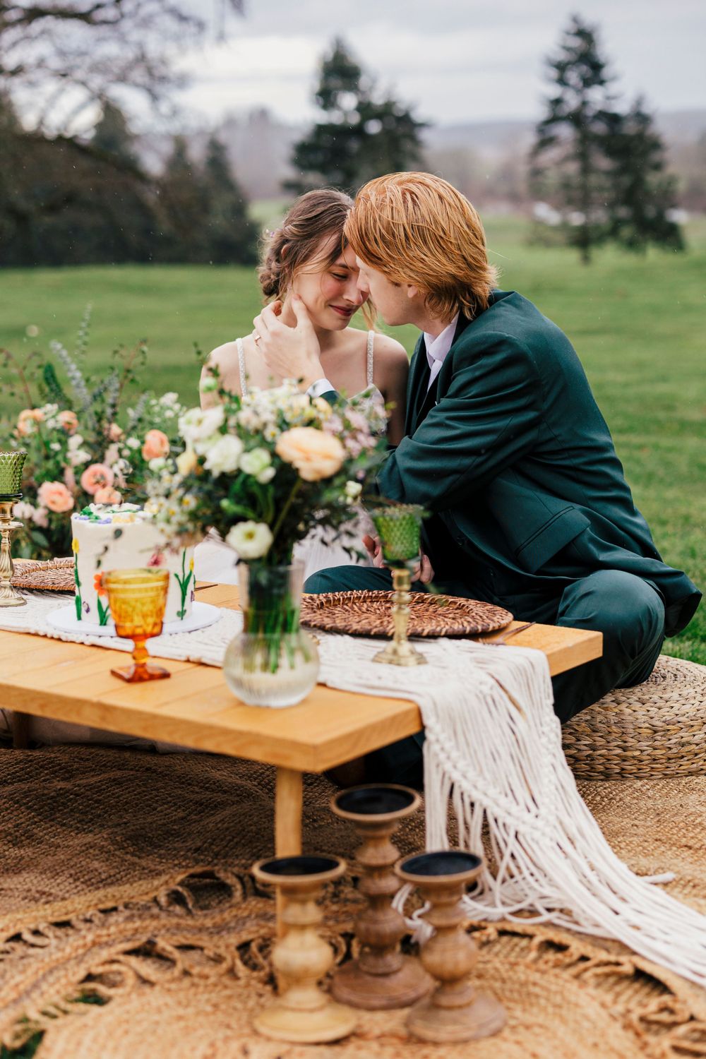 A romantic outdoor wedding setup with velvet green suit, floral arrangements and boho decor on a rustic wooden table.
