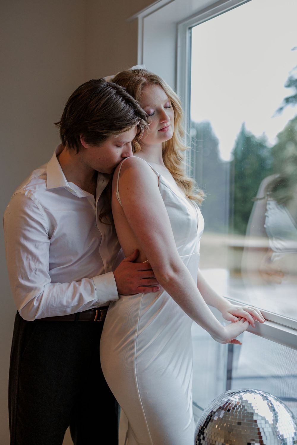 A romantic couple in formal white attire embraces by a window with a disco ball at their feet.