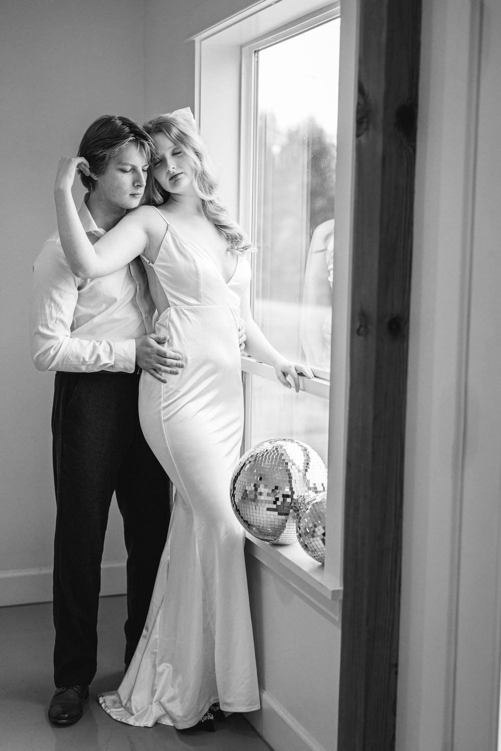 A couple in formal attire embrace by a window with a disco ball, captured in black and white photography.