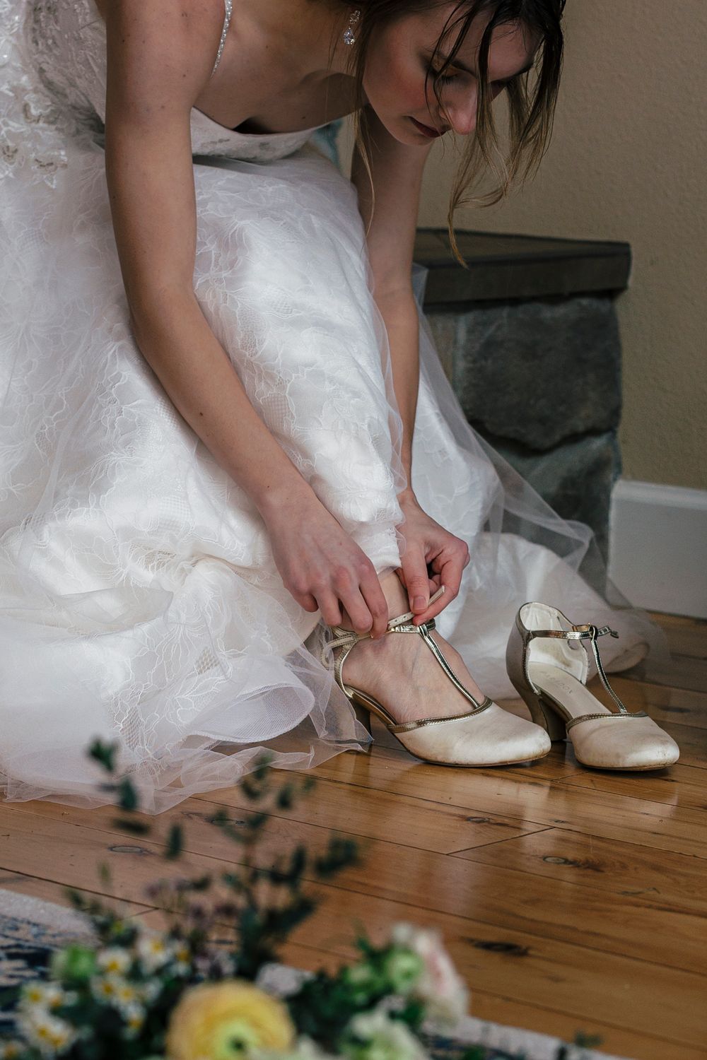 A bride in a white dress fastens her vintage-style shoes while sitting on a wooden floor with flowers nearby.