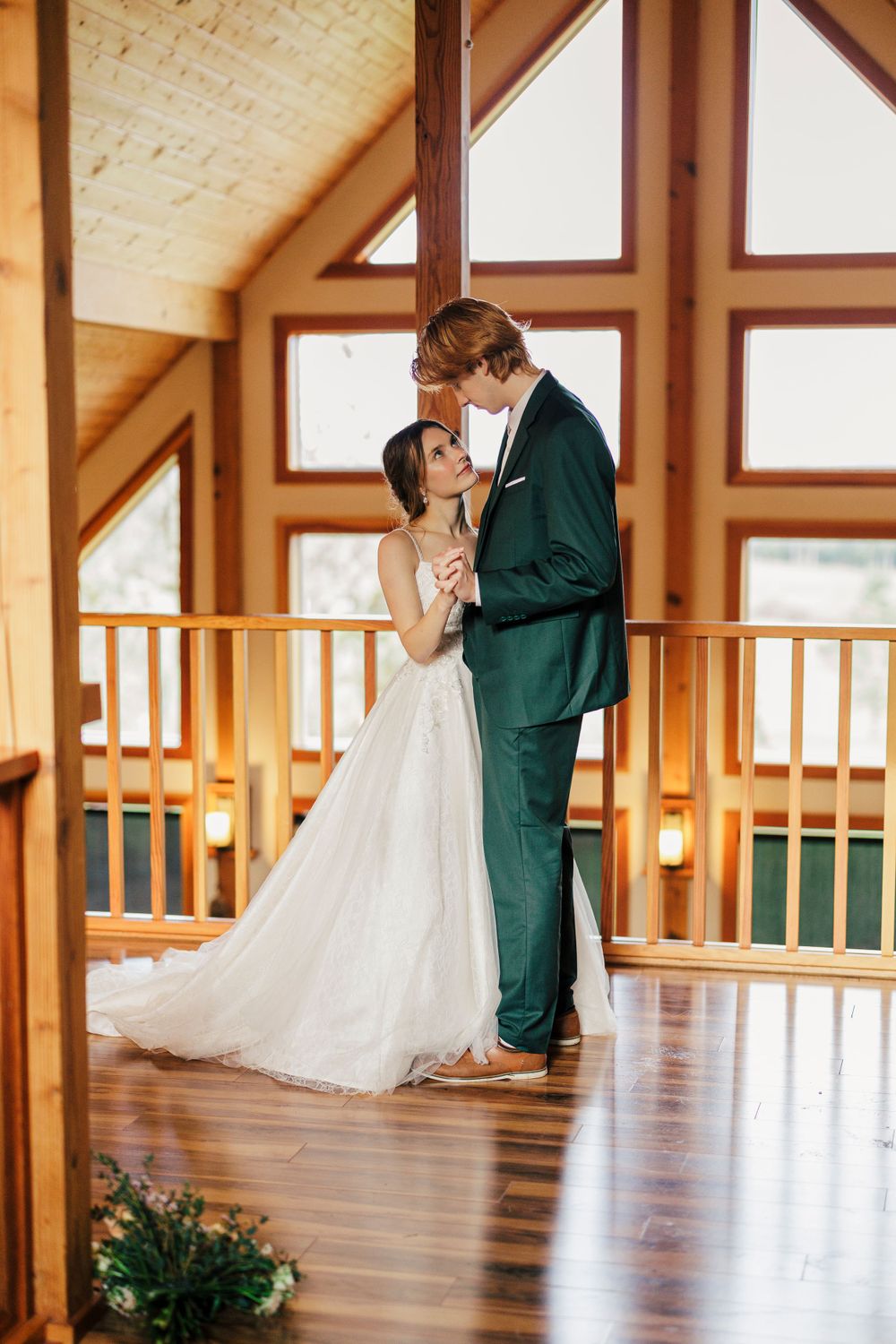 A couple in wedding attire share an intimate moment in a rustic wooden lodge with vaulted ceilings and natural lighting.