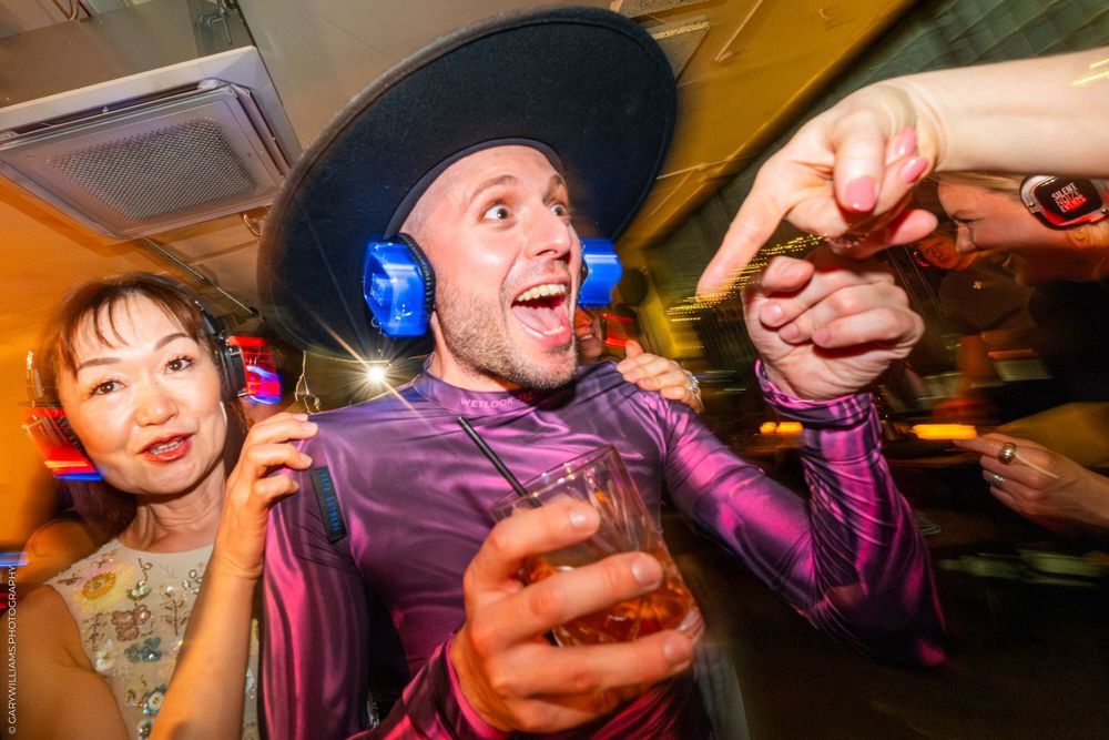 Two people celebrate energetically at a party with one wearing a purple shirt and black hat while holding drinks.