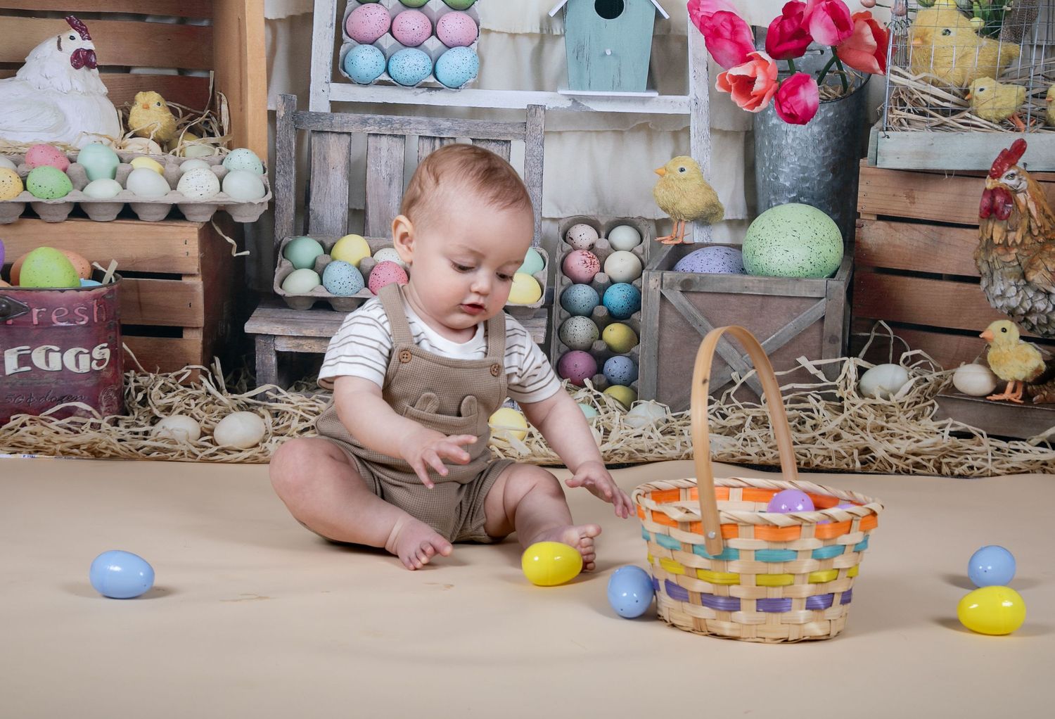 Baby in tan romper sits on floor with colorful Easter basket and eggs in springtime holiday setting.