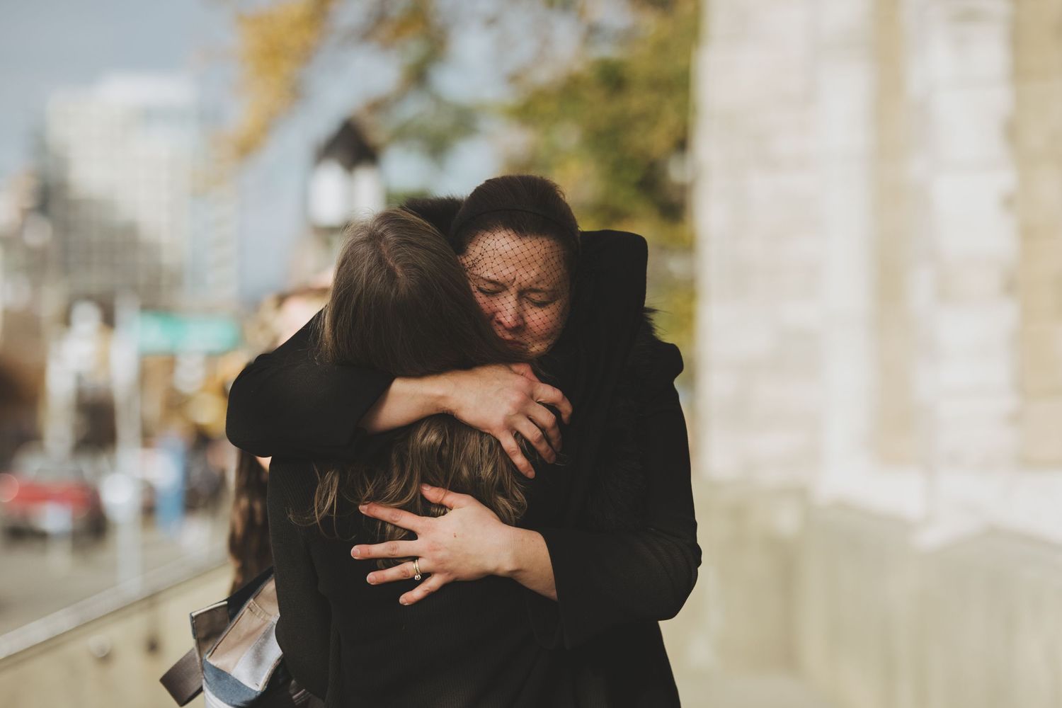 Two people share an emotional embrace on a city sidewalk in warm sunlight.
