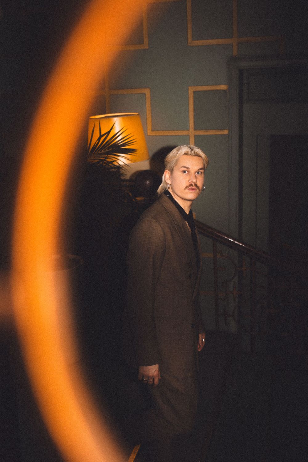 Artistic portrait with orange light flare of person in dark suit against geometric wall pattern.