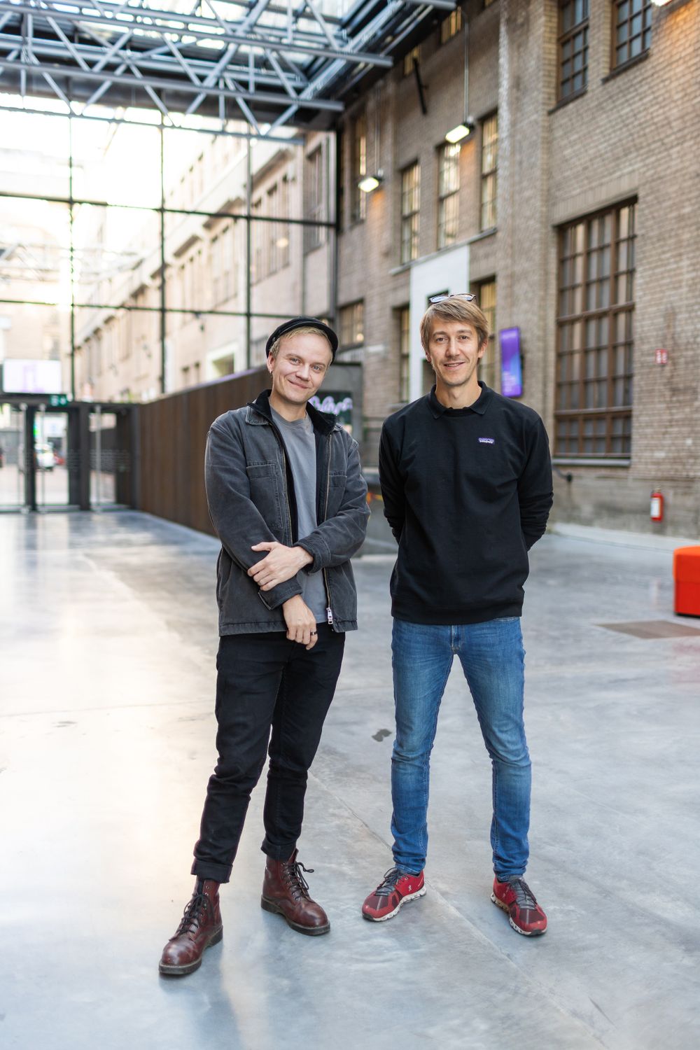 Two people stand casually in industrial building interior with exposed brick walls.