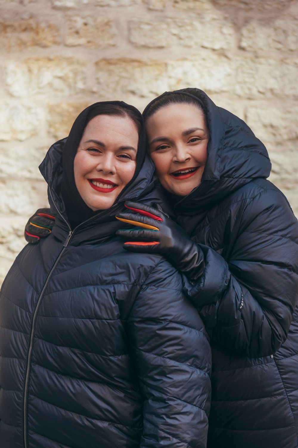 Two people in black puffer jackets smiling together against stone wall.