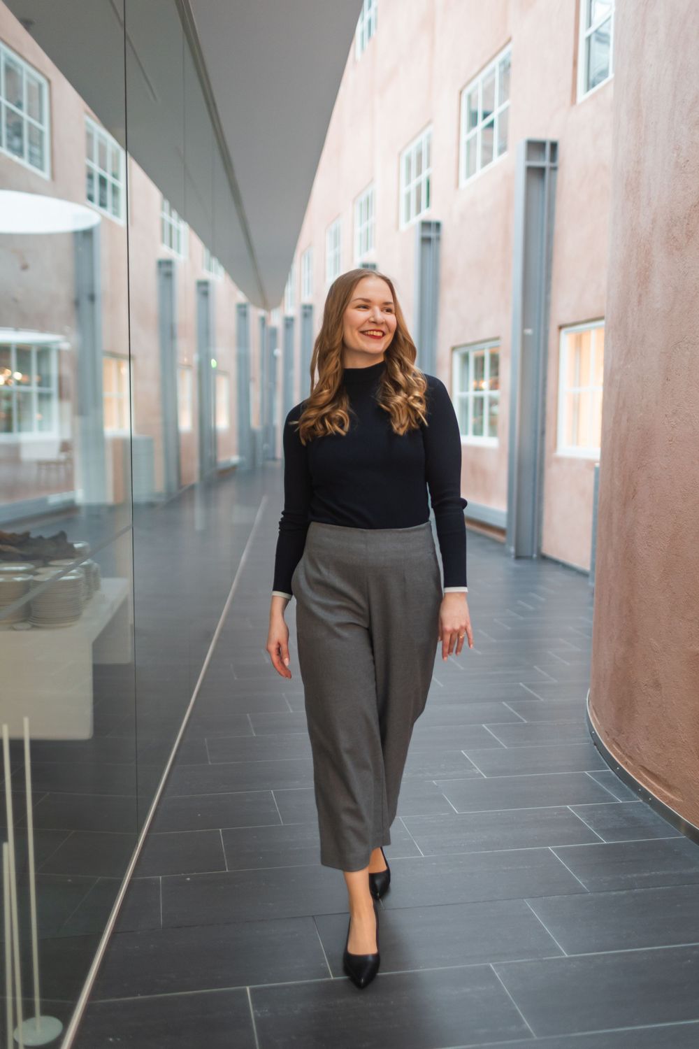 Professional walking confidently down modern hallway wearing black turtleneck and grey pants.