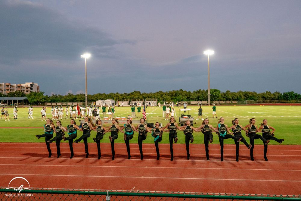 Line of cheerleaders performing synchronized high kicks on a track field at night under stadium lights.