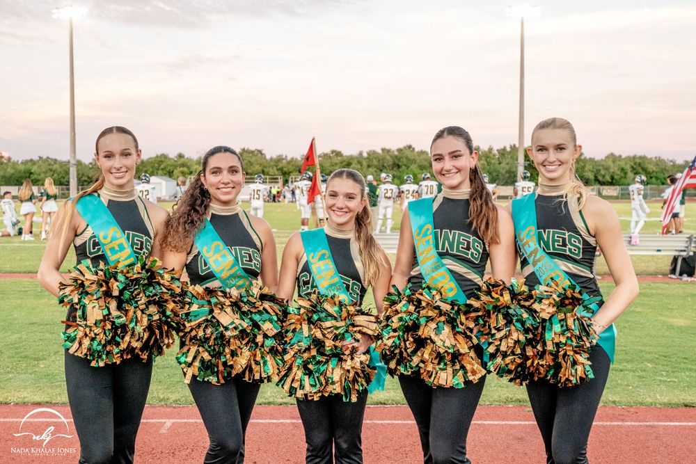 Cheerleaders in green uniforms with orange and black pom poms pose together on a sports field.