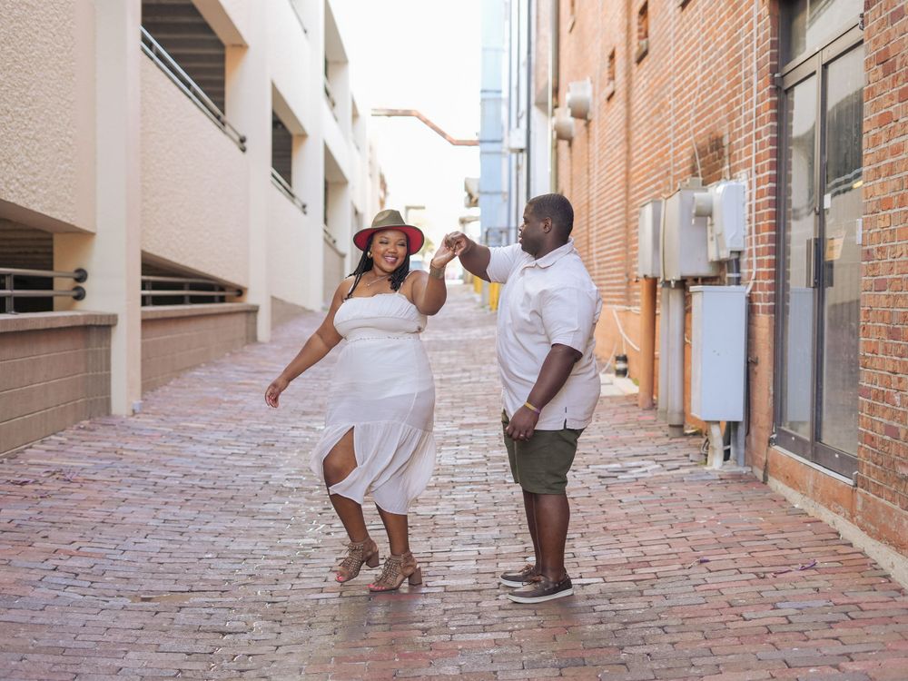 A couple dancing playfully together on a brick paved street between buildings.