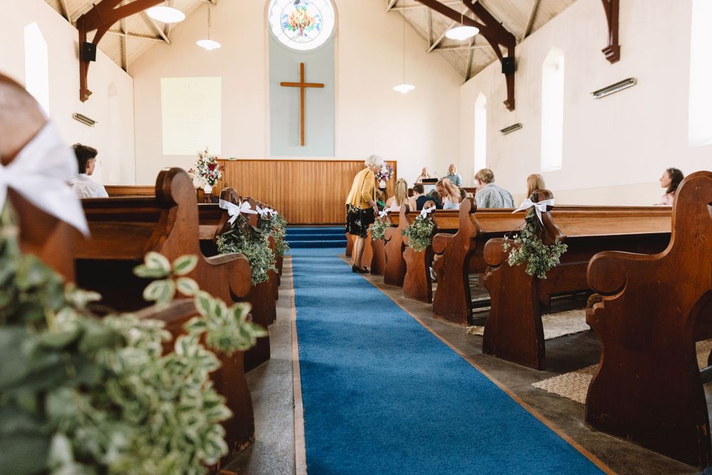 Interior of a small church with wooden pews decorated with floral arrangements and a blue carpet aisle.