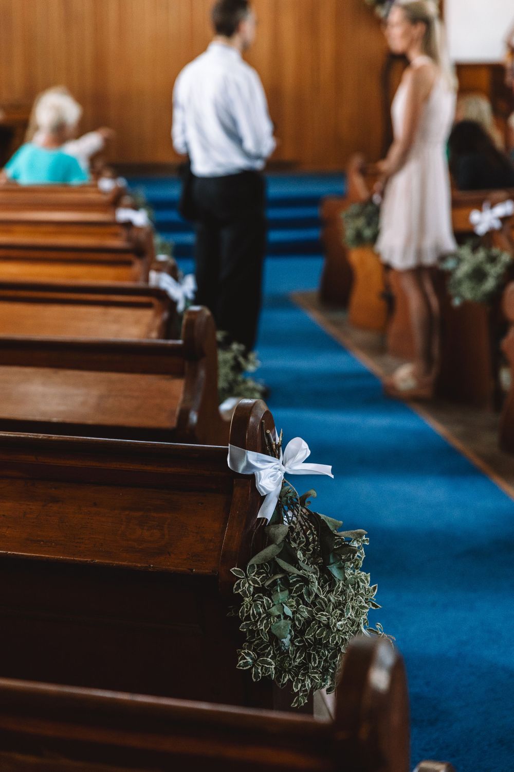 Church pew decorated with greenery and white ribbon along a blue carpeted aisle.