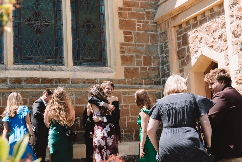 Wedding guests chatting in front of a stone church building on a sunny day.