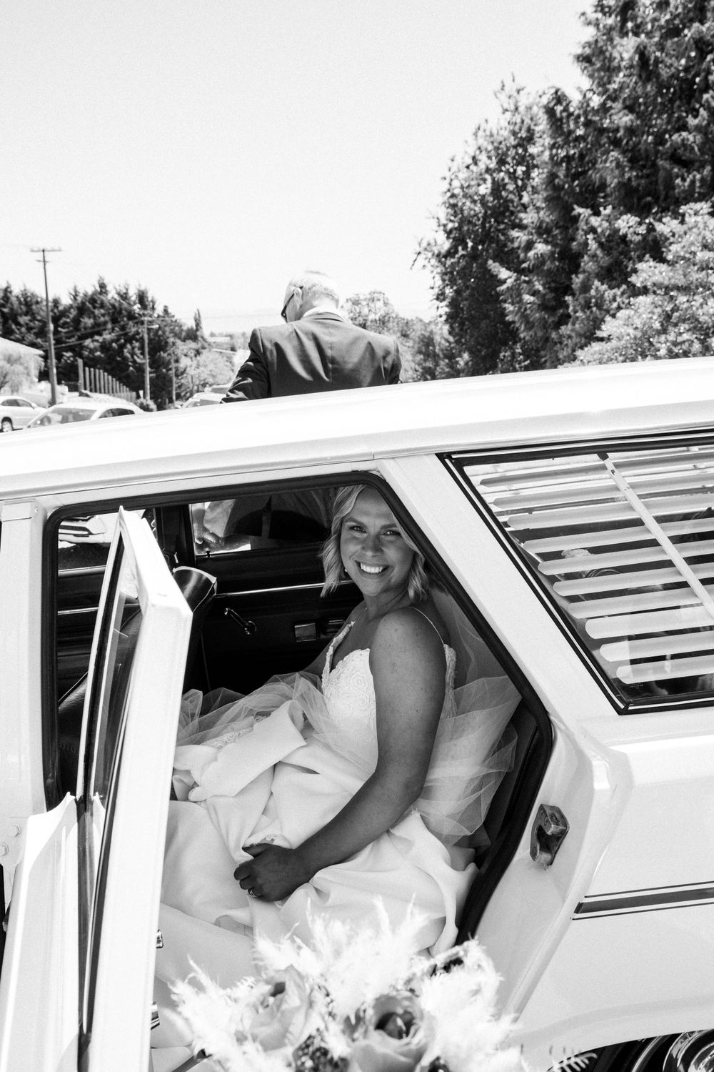 Black and white photo of passenger in wedding dress inside classic car.