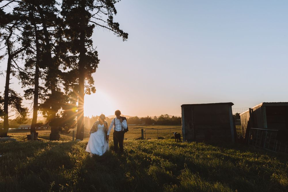 Wedding couple walks through rural field at sunset with rustic barn buildings in background.