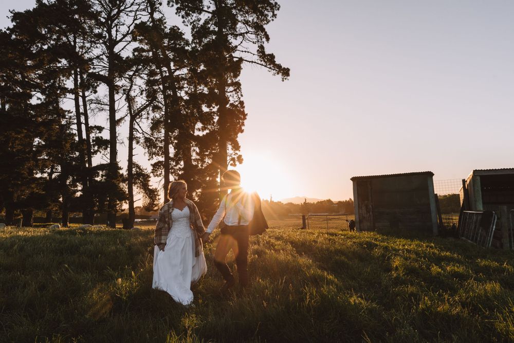 Newlyweds stroll hand in hand through grassy meadow during golden hour near farm buildings.