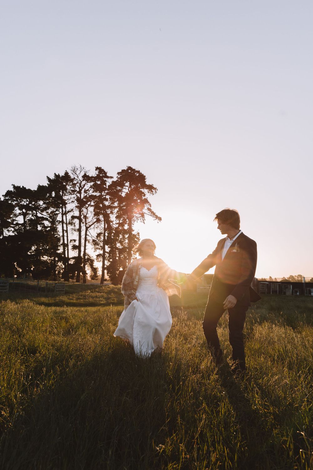 Couple dances playfully in grassy field during sunset with pine trees silhouetted in background.