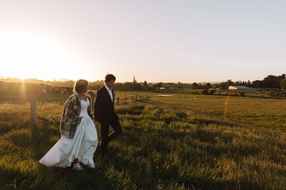 Wedding couple walks through pastoral field during sunset with rustic farm buildings in distance.
