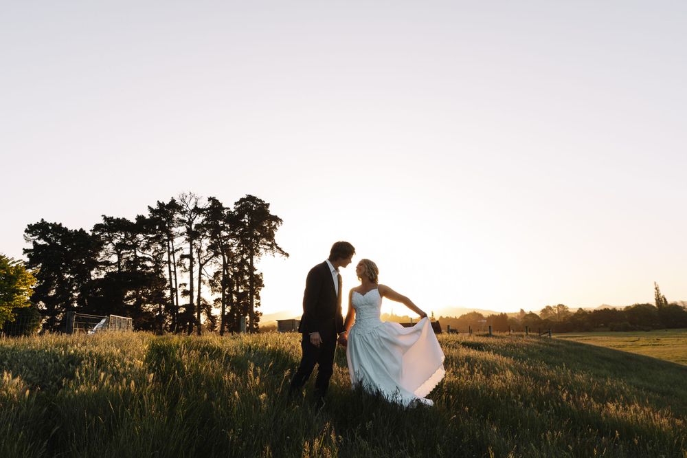 Wedding couple silhouetted against sunset sky on grassy hilltop with pine trees.