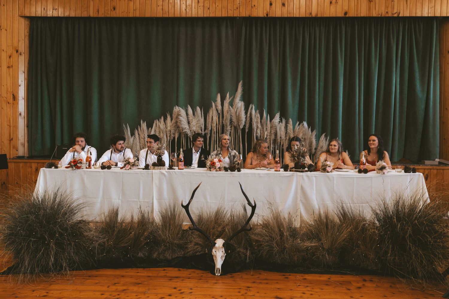 Wedding reception head table decorated with pampas grass and antler decor in rustic wooden venue.