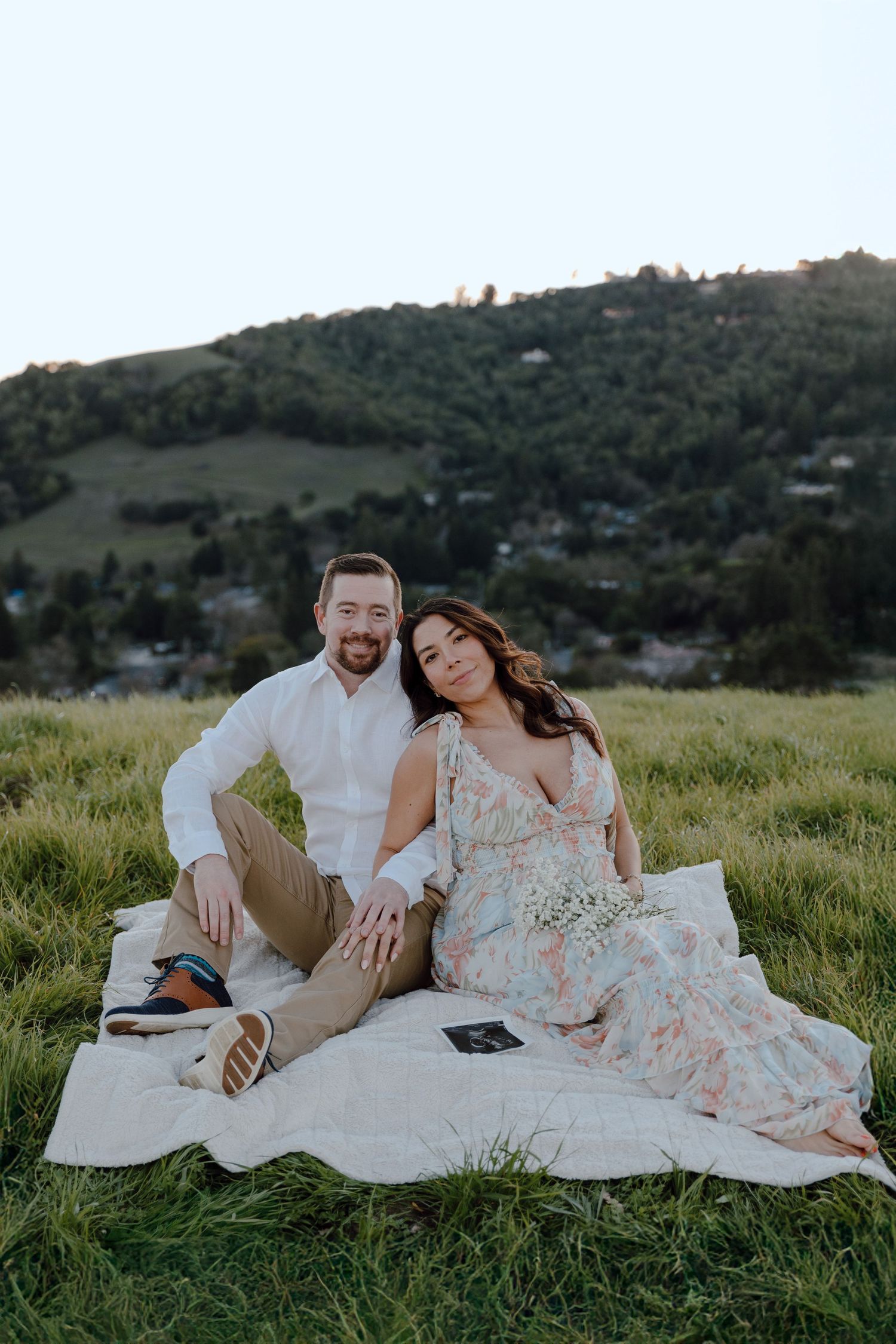 A couple sits on a blanket in a grassy hillside setting during golden hour.