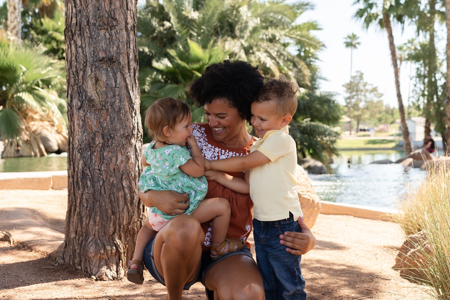 Three people sharing a tender moment together on a bench near a tranquil pond with palm trees.