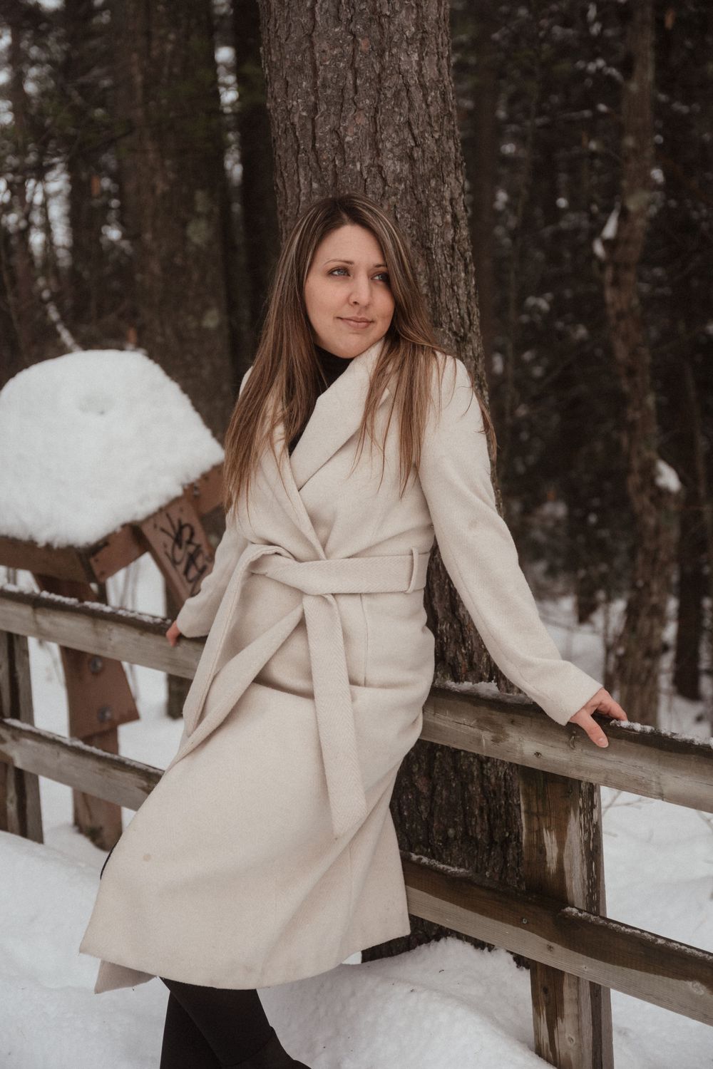 Woman in a cream coat poses on a wooden bridge in a snowy forest landscape.