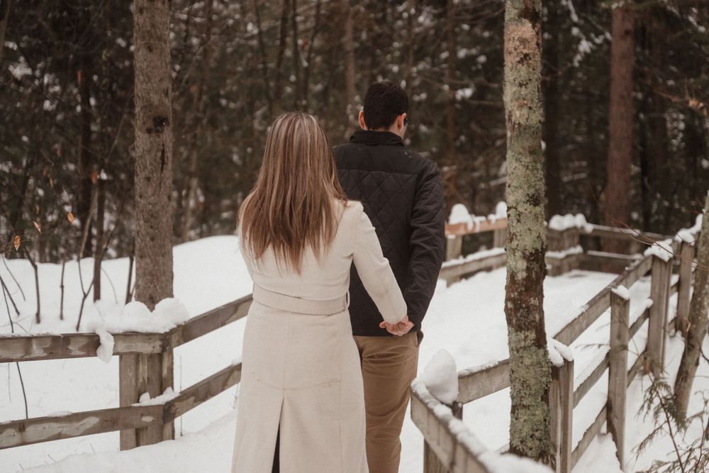Couple walking hand in hand on a wooden boardwalk through a snow-covered forest.