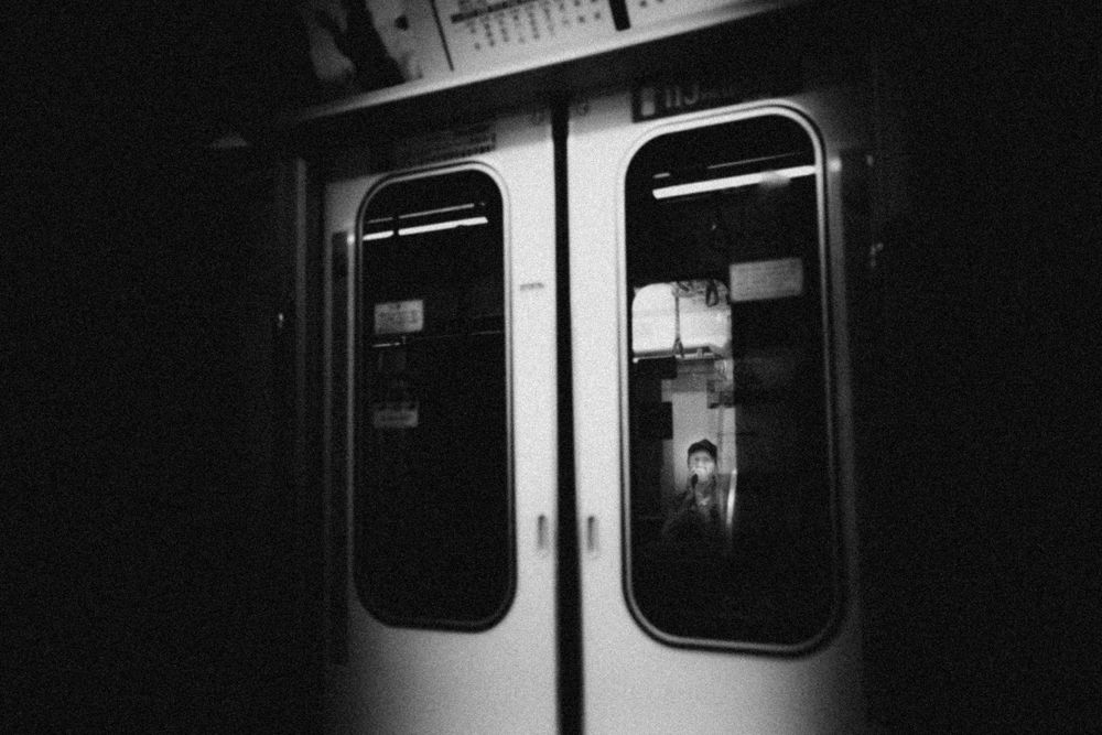 A dark, moody black and white photograph showing a subway car door window at night.