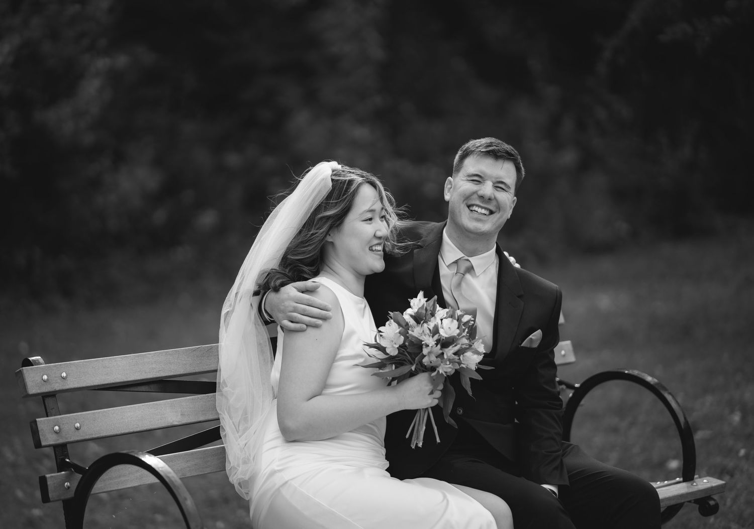 Black and white wedding portrait of couple sharing laughter on park bench.