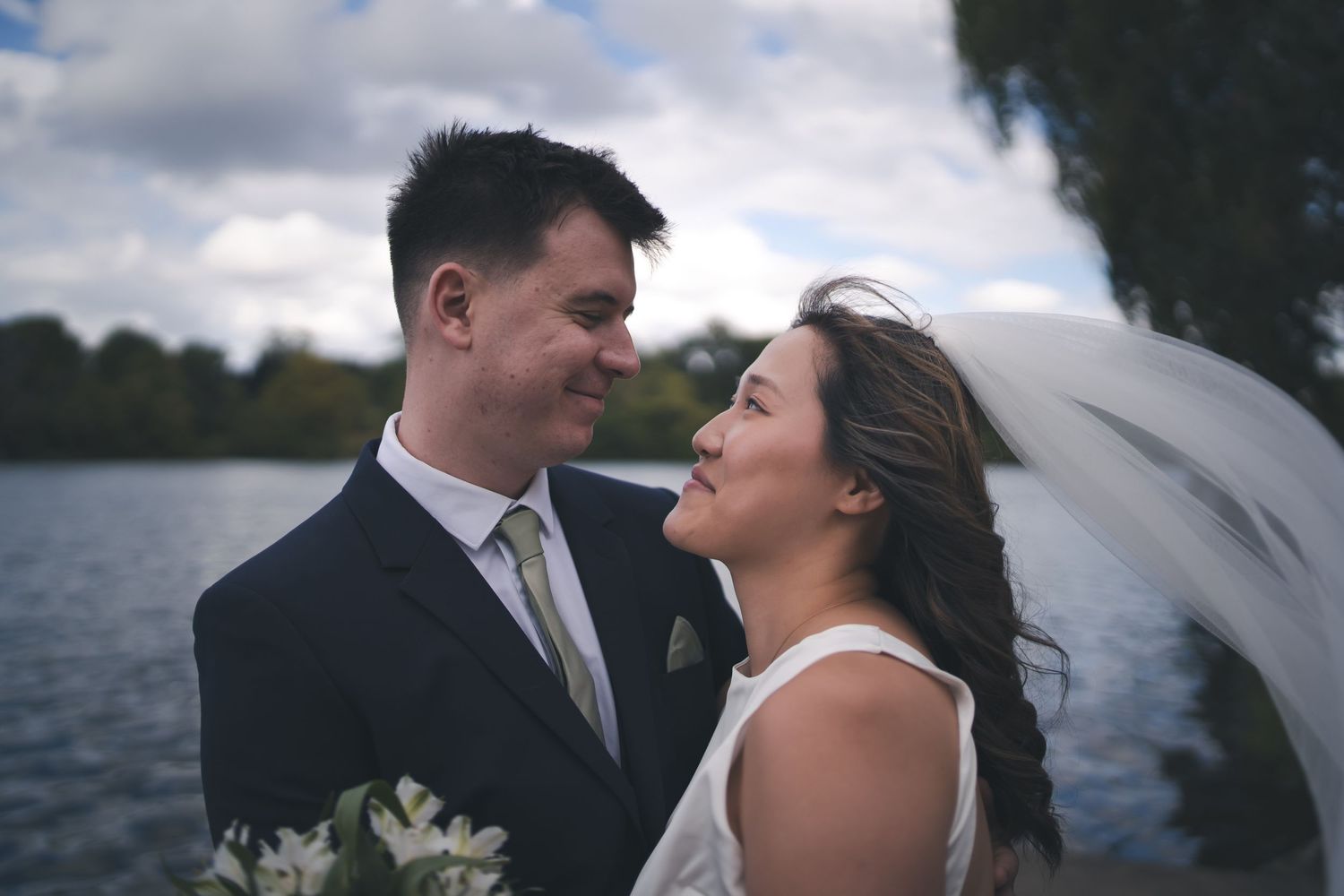 Intimate wedding portrait by lakeside with flowing veil in breeze.