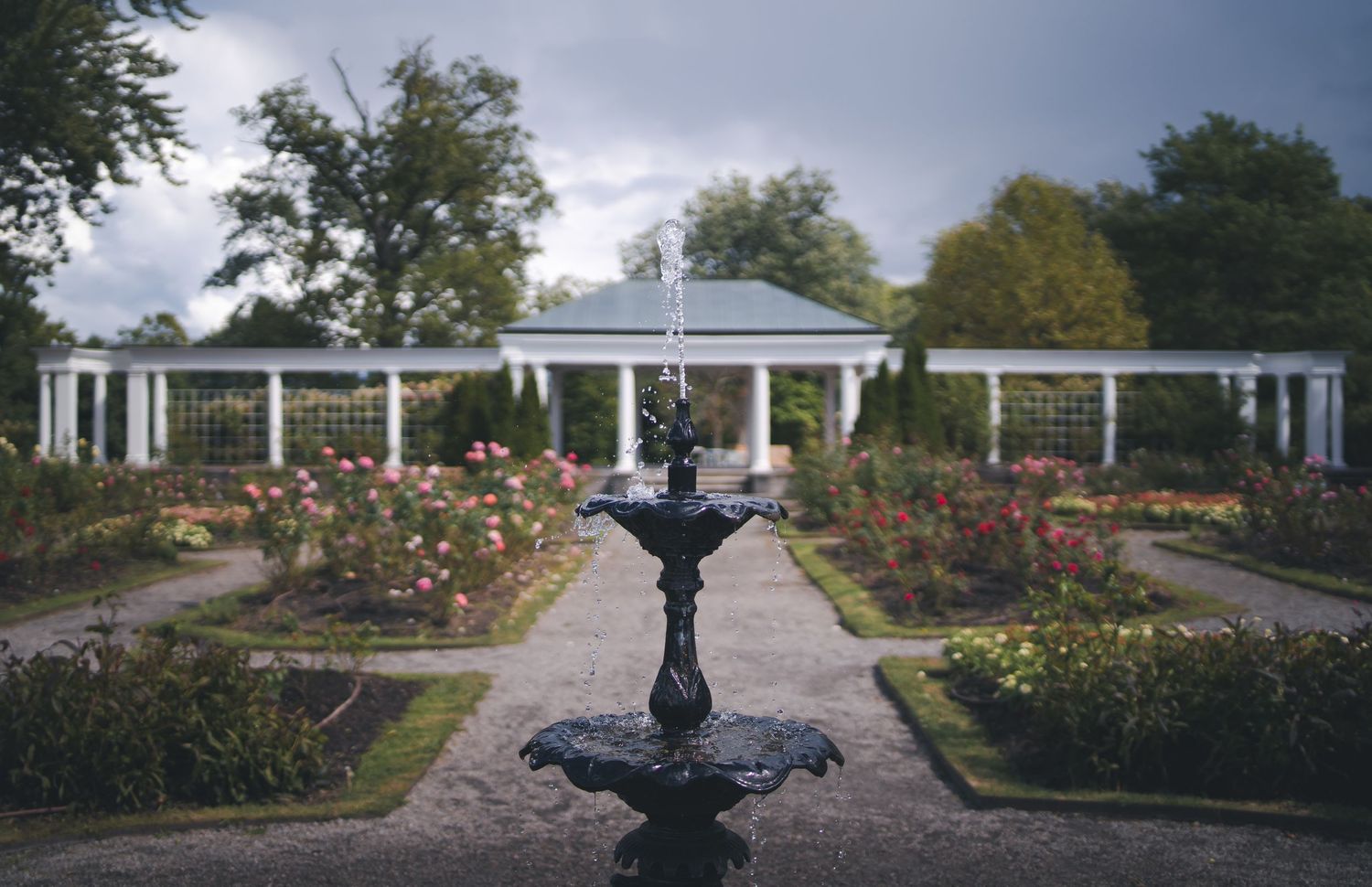 A formal garden features a decorative fountain surrounded by white pergolas and blooming flowers.