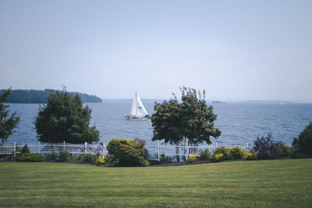 A sailboat glides across a lake viewed through trees from a manicured waterfront lawn.